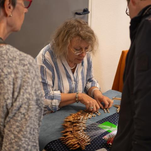 Vrouw demonstreert kantklossen aan twee geïnteresseerde bezoekers die over haar tafel staan gebogen.