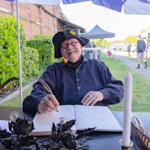 Man met hoed, bril en pijp demonstreert het schrijven met een ganzenveer.